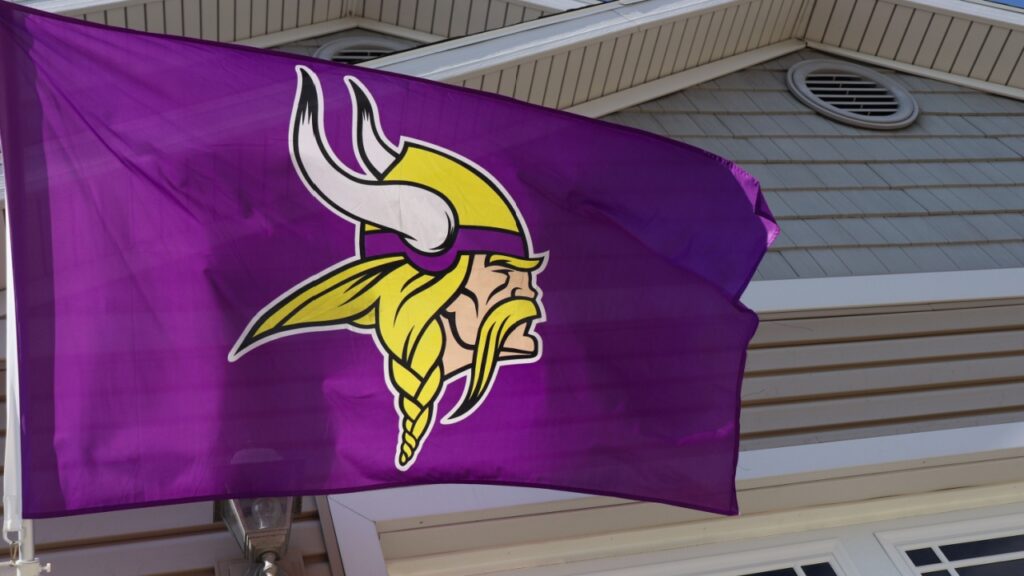 A purple-and-gold Minnesota Vikings flag waves in the wind against a house. 
