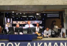Troy Aikman in a broadcasting booth with commentators and equipment during a sporting event.
