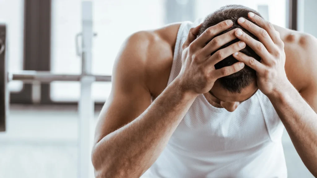 Upset sportsman touching head while sitting near barbell in gym. 