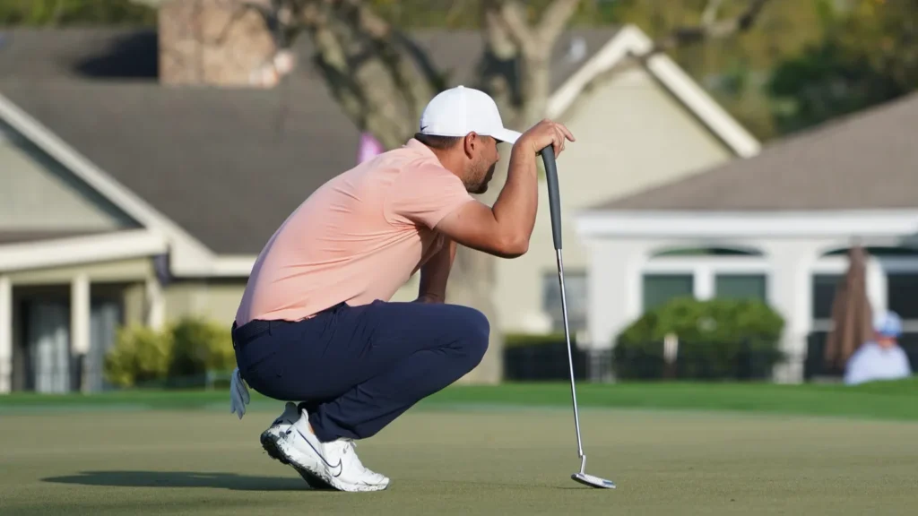 A golfer reading a putt on the green, holding his putter.​