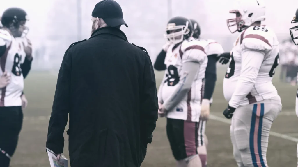 Head coach in black apparel discussing play with the football team on the sideline.