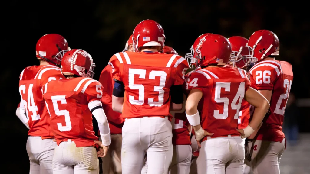 Professional football players in red jerseys huddle together on the field at night.
