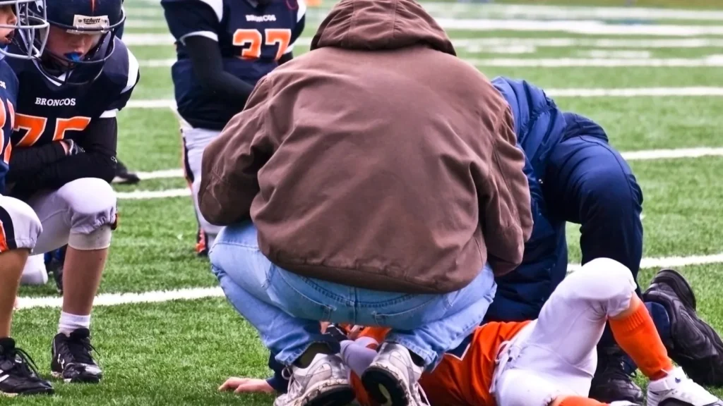 A football player in a uniform lying on the field while coaches kneel to check on him during a game.