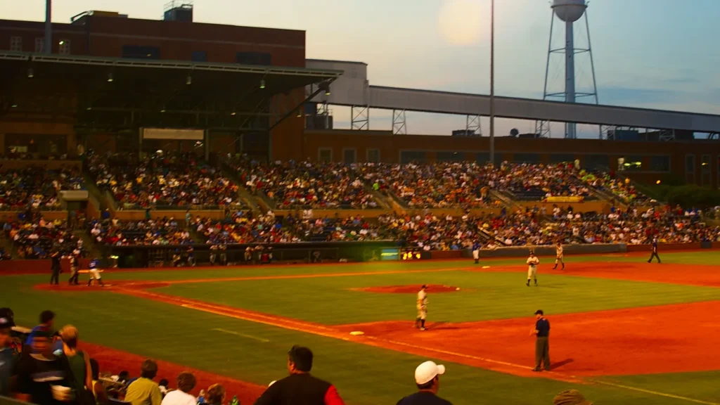 A packed baseball stadium at dusk with players on the field and fans watching under bright stadium lights.