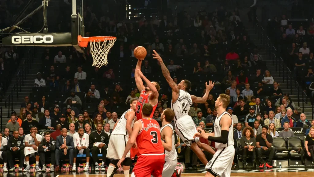 Basketball player in red jersey shoots over defender during professional game with crowd watching.