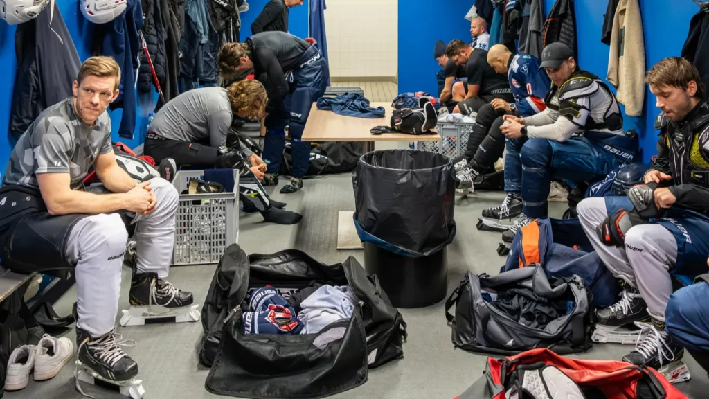 Ice hockey players in full gear sitting inside a team locker room.