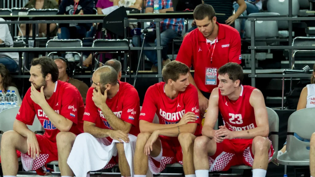 Basketball players on bench watching game with towels and water bottles.