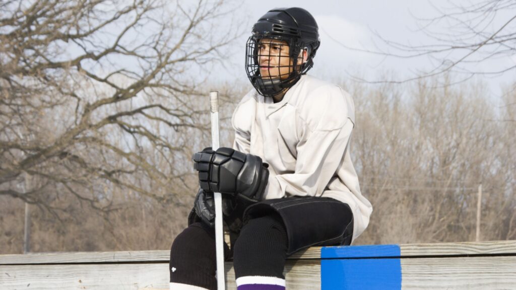 An ice hockey player holding a hockey stick sitting on the sideline.