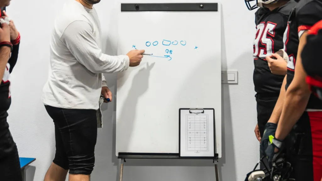 American football coach discussing game strategy on a whiteboard with his team in the locker room planning for success