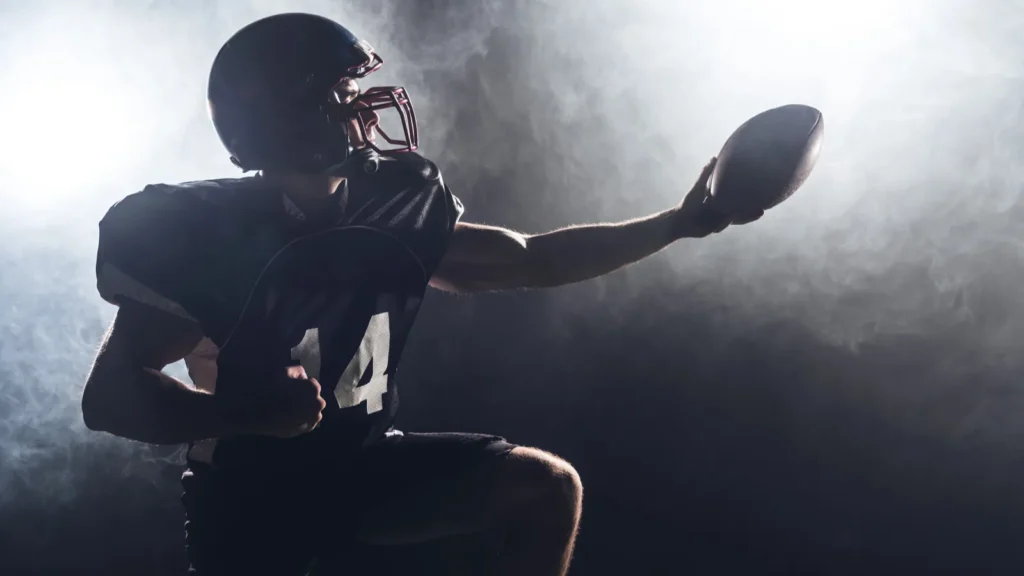 American football player standing on knee on green grass and holding ball against white smoke