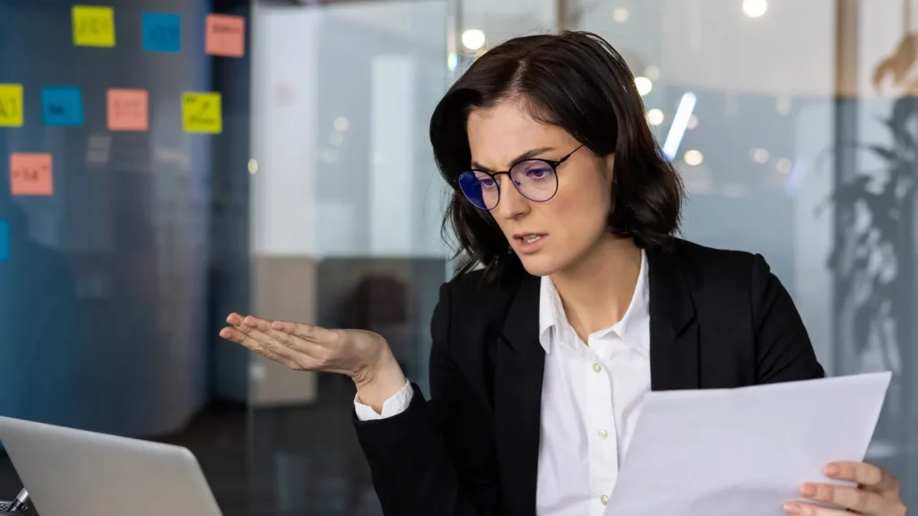 Businesswoman looks confused while analyzing financial documents at laptop in modern office Concept of stress finance and decisionmaking highlighted in workplace setting