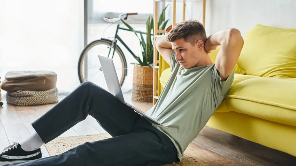 confused man on floor near yellow couch at home studying in laptop and putting hands behind head