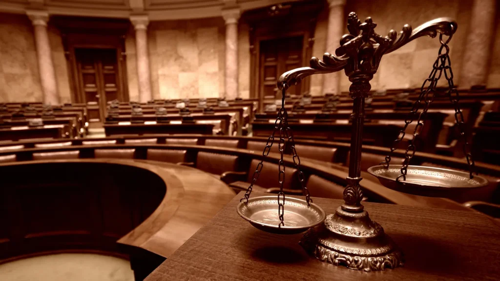 Antique scales of justice in the foreground of an empty legislative chamber.