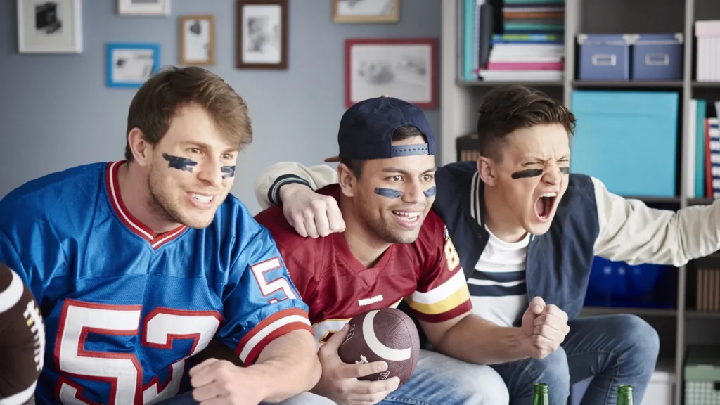 Three football fans cheering while watching a game, holding a football on a couch.
