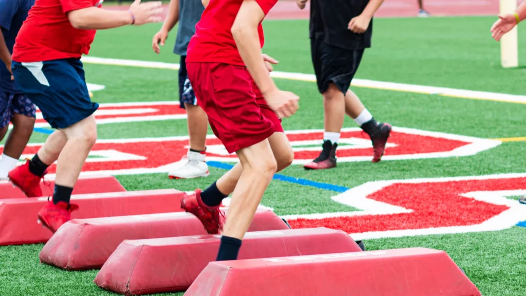 High school football players at summer camp running sideways over red barriers while the coaches watch them at summer training camp