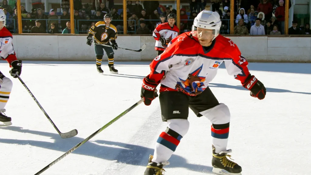 Hockey players competing in an outdoor rink with a crowd of spectators in the background.