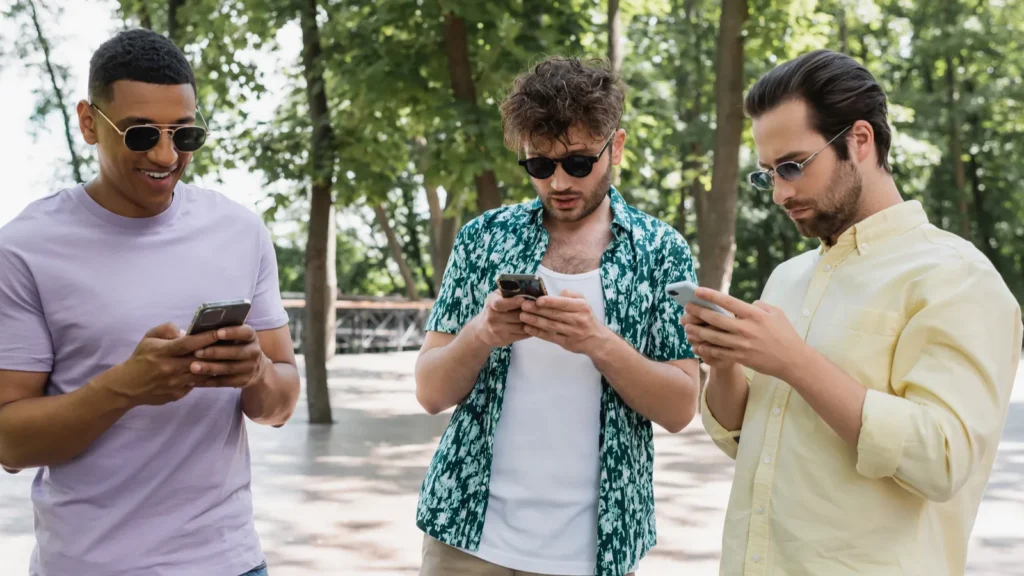 cheerful african american man in sunglasses using mobile phone with stylish friends in city park
