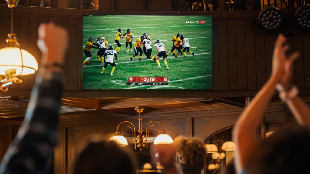 American football fans cheer while watching a live match broadcast in a sports pub.