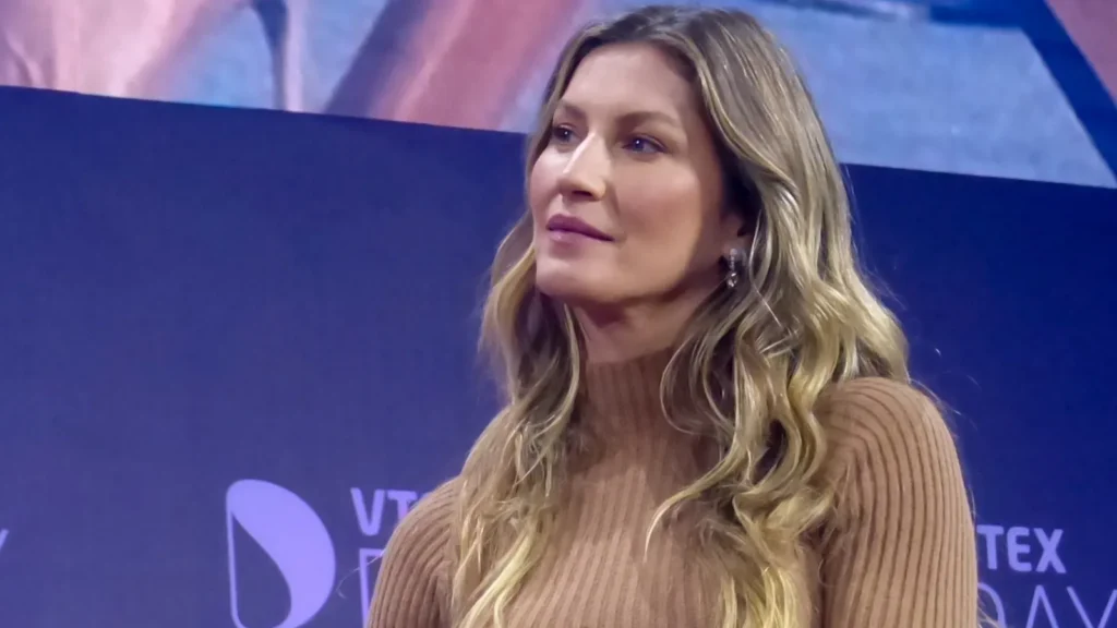 This image shows Gisele Bündchen wearing a ribbed beige sweater at the VTEX Day event in São Paulo, Brazil