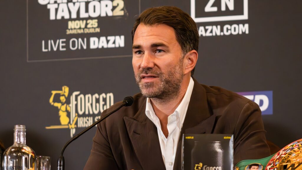 Eddie Hearn speaking into a microphone at a press conference, with a DAZN-branded backdrop behind him