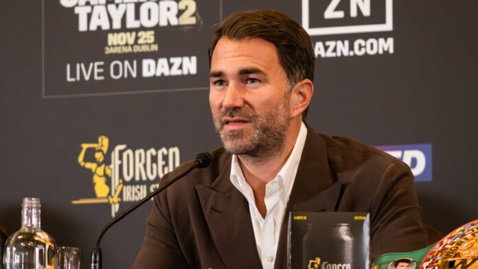 Eddie Hearn speaking into a microphone at a press conference, with a DAZN-branded backdrop behind him