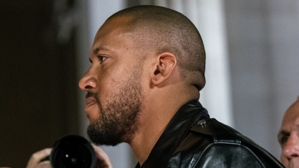 Side profile of a man with a short beard and shaved head wearing a black leather jacket; a photographer’s camera lens appears in the foreground.