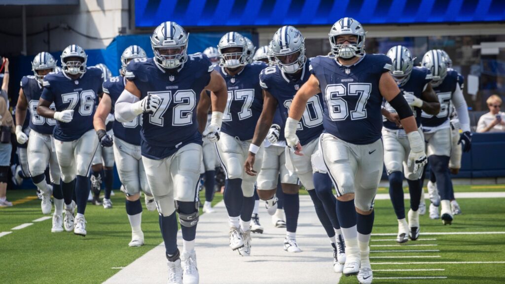 Group of American football players in navy uniforms sprinting onto the field from a tunnel, helmets on, white pants and cleats visible on a green turf.
