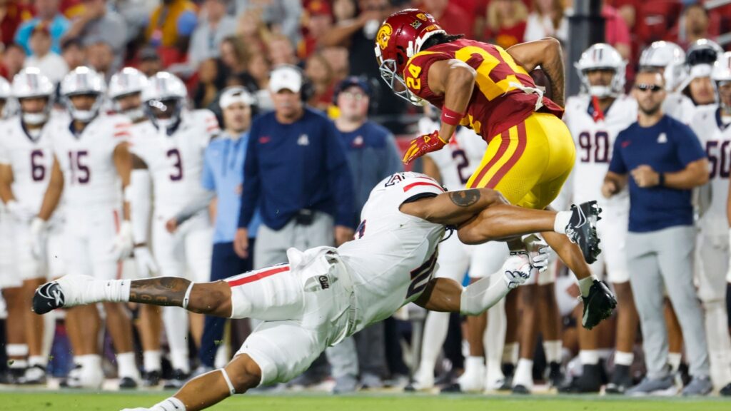 Football action: defender in white tackles a USC player in maroon and gold as he leaps over him near the sideline with a crowded stands backdrop of fans.