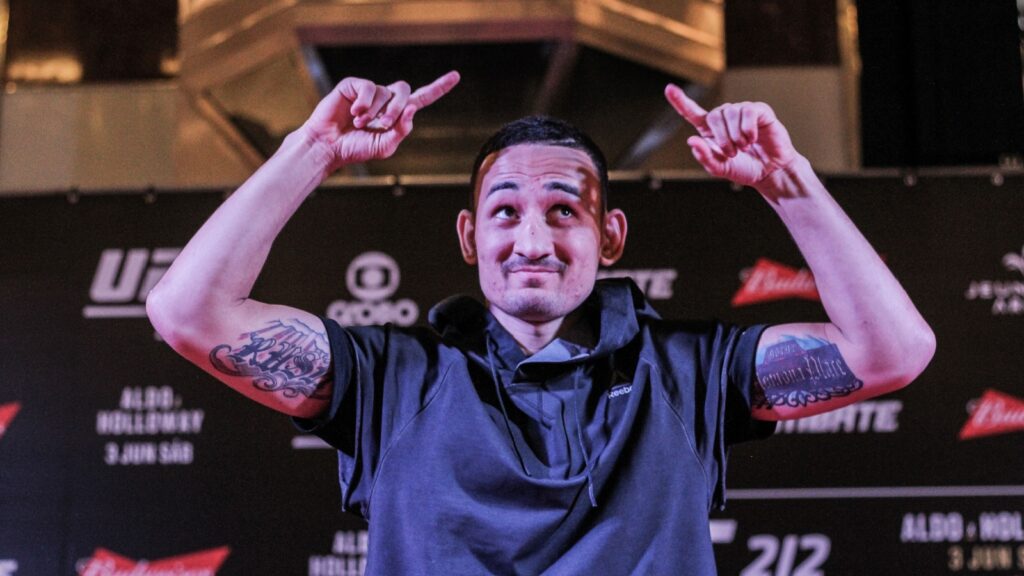 Male fighter on stage, pointing upward with both index fingers, wearing a black hoodie; tattoos on both arms visible.