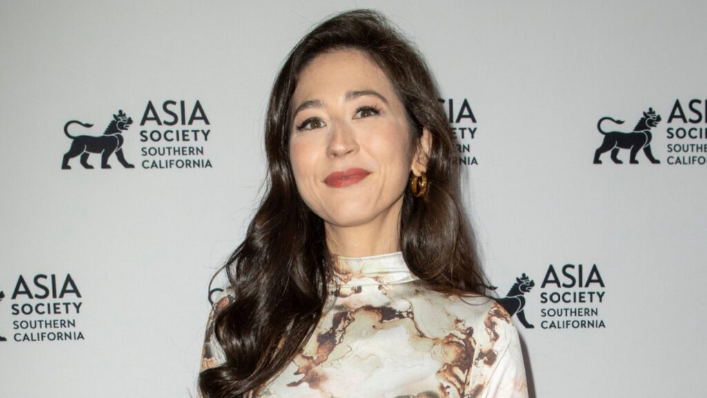 Mina Kimes with long dark hair and a marble-patterned blouse smiles at the camera in front of an Asia Society backdrop.