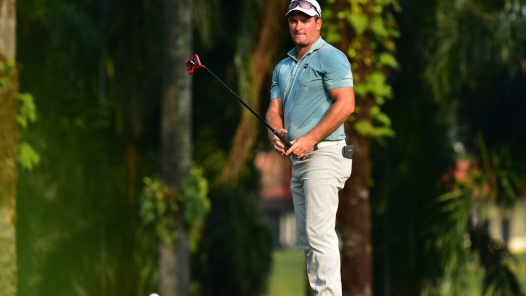 Ryan Fox in a teal polo and white cap holding a golf club on a sunlit course, preparing to swing.