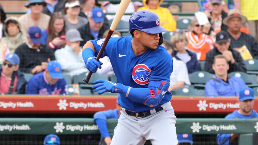 Cubs batter in blue uniform swings at a pitch, crowd watching behind him at a baseball game