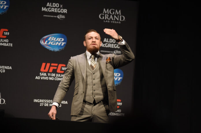 Conor McGregor in a gray pinstripe suit on a stage at a UFC event, raising his right hand