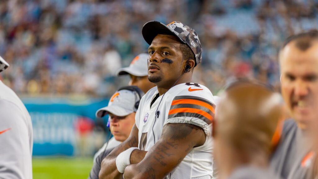 Deshaun Watson in white Cleveland Browns jersey with orange stripes stands with arms crossed on the sideline during a game, stadium crowd in the background.