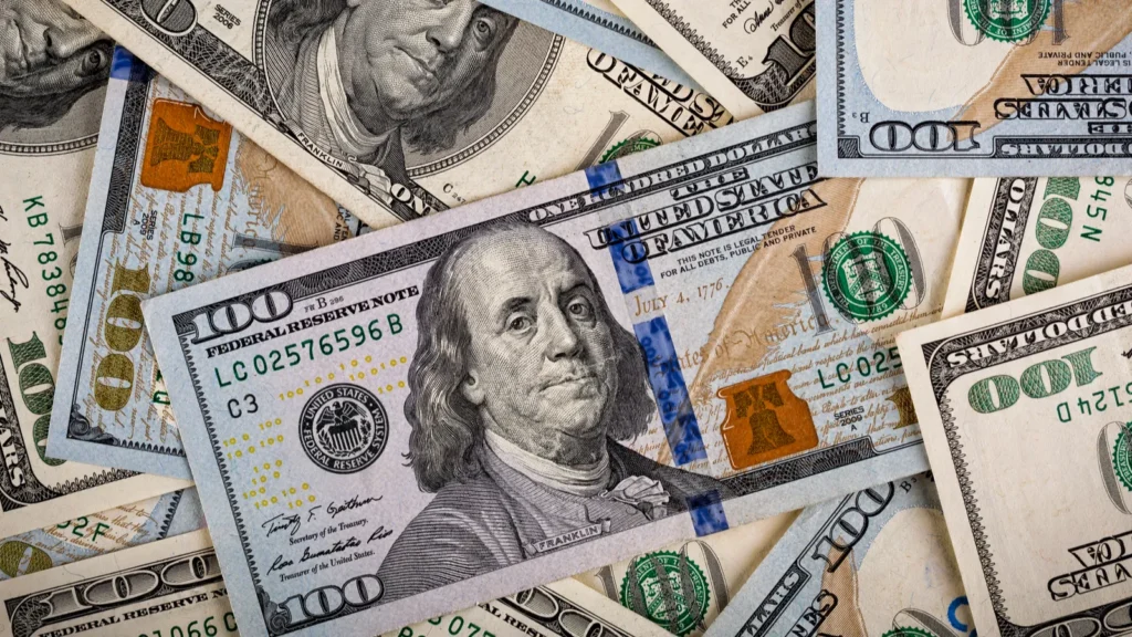 Pile of overlapping U.S. hundred-dollar bills, Benjamin Franklin visible with blue security strip, in a money collage.