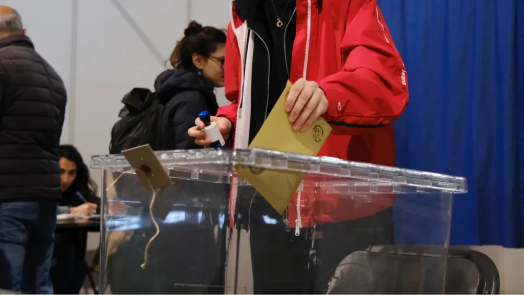 Voter in a red jacket inserts a ballot into a clear ballot box at a polling station, envelope in hand.