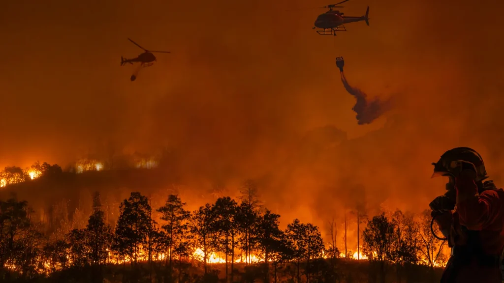 Two firefighting helicopters drop water over a raging wildfire at night.