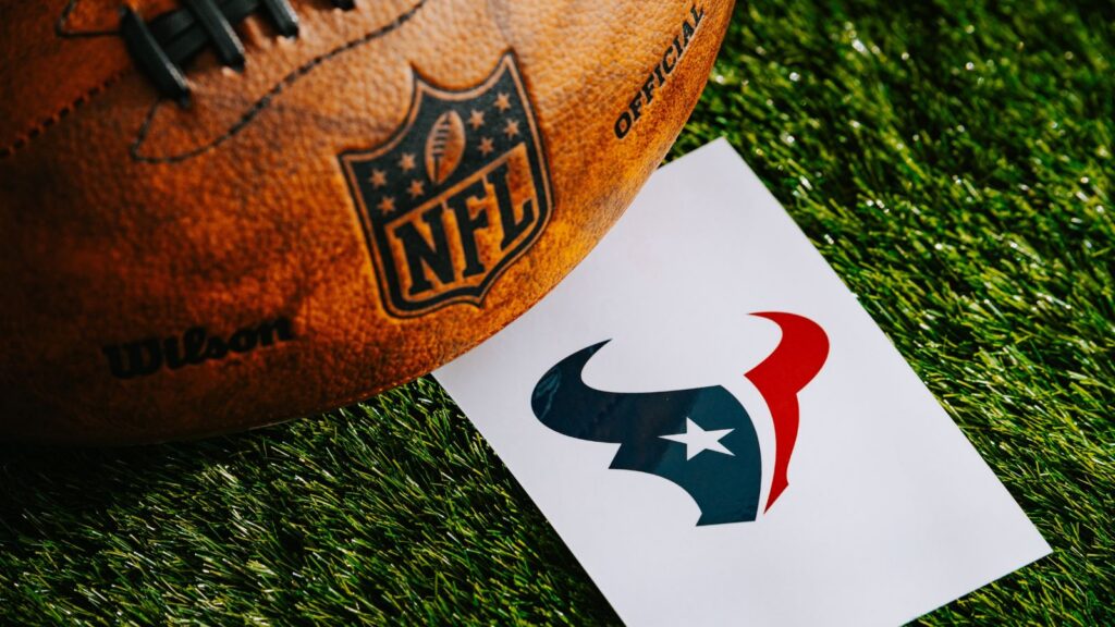 Close-up of an orange NFL football on grass beside a white card featuring the Houston Texans logo
