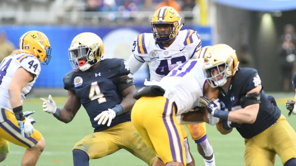 Notre Dame defenders in dark blue jerseys tackle an LSU offensive player during a college football game.