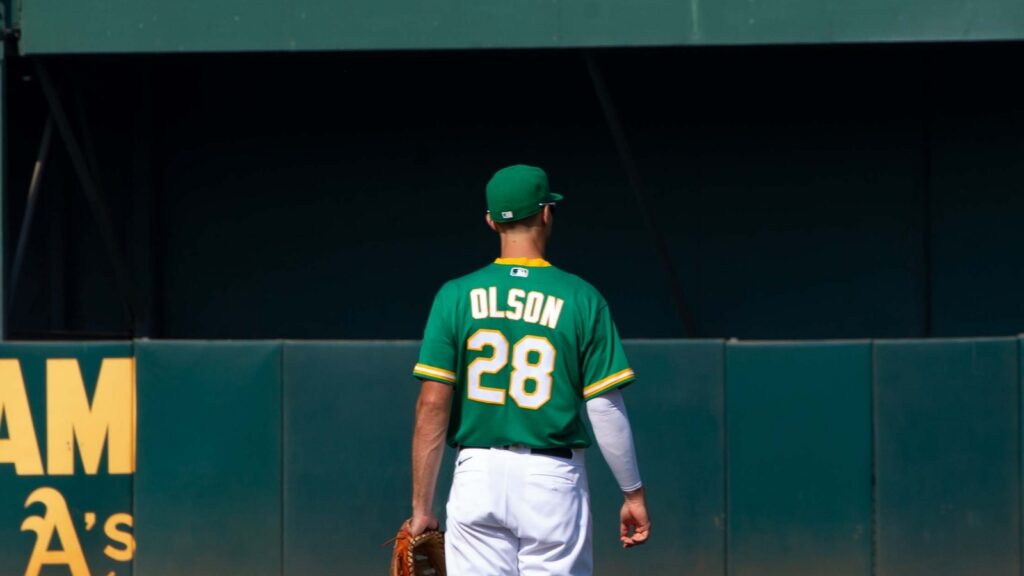 Baseball player with back to camera wearing green Athletics jersey #28 Olson, holding a glove, standing near the outfield wall.