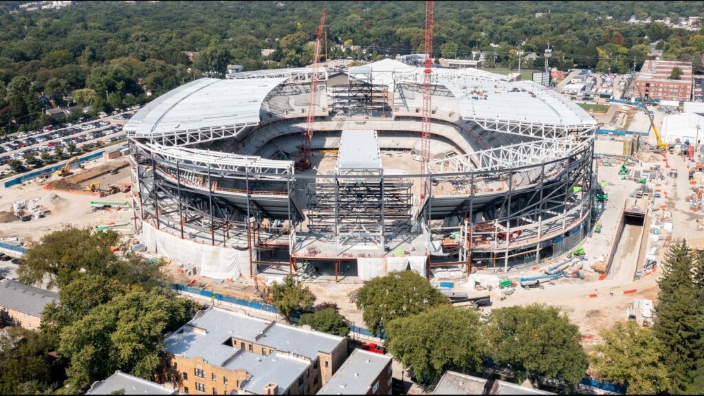 A large stadium under construction with steel framework and two red cranes over the open field center.