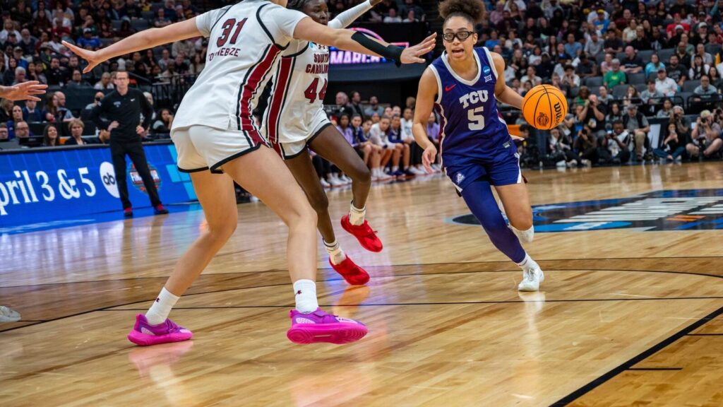 TCU player in blue (#5) dribbles past two white-jersey defenders during a college basketball game.