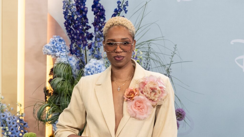 Person wearing a beige blazer with a large pink flower brooch, sunglasses, and short blond hair, posing among blue and purple flowers in a studio backdrop.