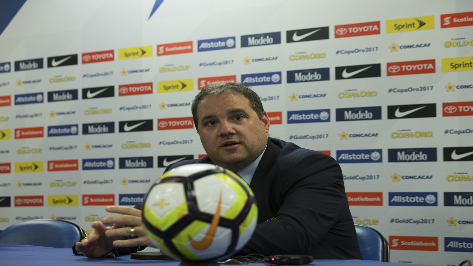 Man in suit at a press conference with a neon soccer ball in the foreground and sponsor logos on the backdrop behind him, speaking to the press.