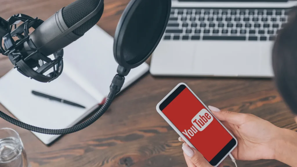 Podcast recording setup with a professional microphone, pop filter, notebook, laptop, and a smartphone displaying the YouTube logo