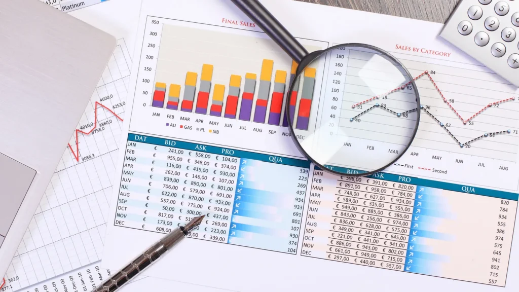 Financial sales reports, charts, magnifying glass, pen, and laptop on a wooden desk.