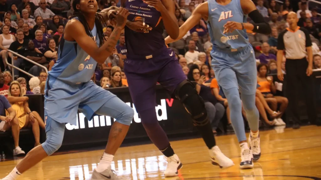 Two Atlanta Dream basketball players in light blue uniforms defend against a Phoenix Mercury player driving on the court.