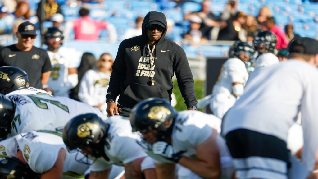 Coach Deion Sanders wearing a black hoodie watches over his football team during practice.