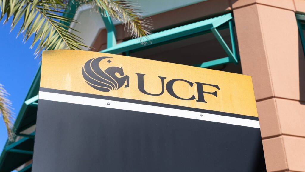 An outdoor campus sign featuring the University of Central Florida (UCF) logo and text against a building and palm tree backdrop.